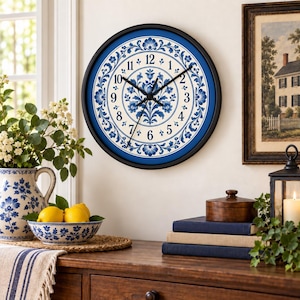 May include: A round wall clock with a black frame and a white face featuring a blue floral pattern. The clock has black hour, minute, and second hands, and black Arabic numerals from 1 to 12 arranged in a circle around the central floral design.