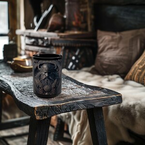 May include: A black ceramic mug with a detailed image of a Viking warrior with a beard and horned helmet. The mug is sitting on a wooden table.