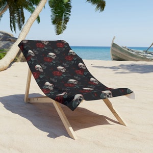 May include: A black beach chair with a skull and rose pattern. The chair is made of wood and has a fabric seat and back. The chair is on a sandy beach with a palm tree in the background.