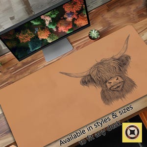 May include: A brown desk mat with a black and white illustration of a Highland cow. The text "Available in styles & sizes to fit any desk" is printed on the mat.