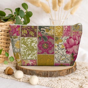 Puede incluir: Un neceser rectangular con diseño de patchwork. La bolsa presenta varios estampados florales en tonos rosa, verde y dorado. Una gran peonía rosa es visible en el lado derecho. Tiene una cremallera dorada y un interior blanco.