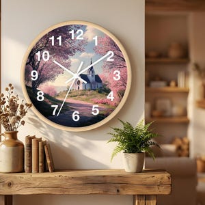 Könnte beinhalten: Runde Wanduhr mit hellem Holzrahmen. Das Zifferblatt zeigt ein szenisches Bild einer Kirche auf einem Weg, der von rosa blühenden Bäumen umgeben ist. Weiße Zeiger und Ziffern vervollständigen das Design.