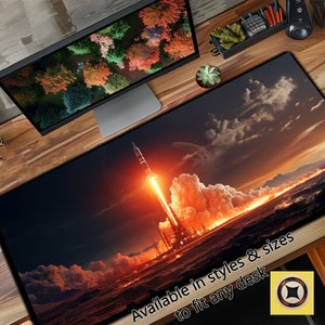 May include: A dark blue desk mat with a rocket launching into space with a fiery orange trail. The text "Available in styles & sizes to fit any desk" is at the bottom of the mat.