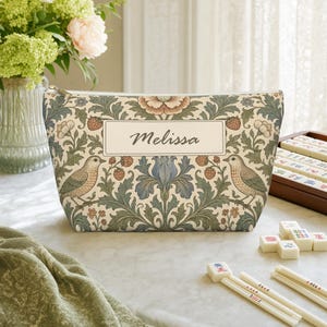 May include: A zipped pouch featuring a cream-coloured background with a botanical pattern of green leaves, blue flowers, red strawberries, and two birds. A central rectangular label displays the name Melissa in a dark script font.