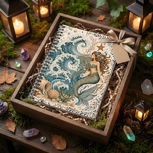 May include: A spiral-bound notebook featuring a mermaid illustration with blue ocean waves, seashells, and a starfish, presented in a wooden gift box filled with shredded paper, moss, crystals, and lanterns with lit candles and a beige ribbon.