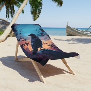 May include: Beach towel featuring a detailed illustration of a black raven perched on a rock, set against a sunset sky with trees. The towel is draped over a wooden beach chair on a sandy beach.