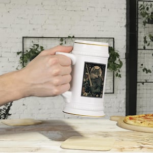 Pode incluir: Caneca de cerveja de cerâmica branca com borda dourada e uma ilustração em preto e branco de um lobo segurando uma espada. O lobo está rodeado de flores e folhas.