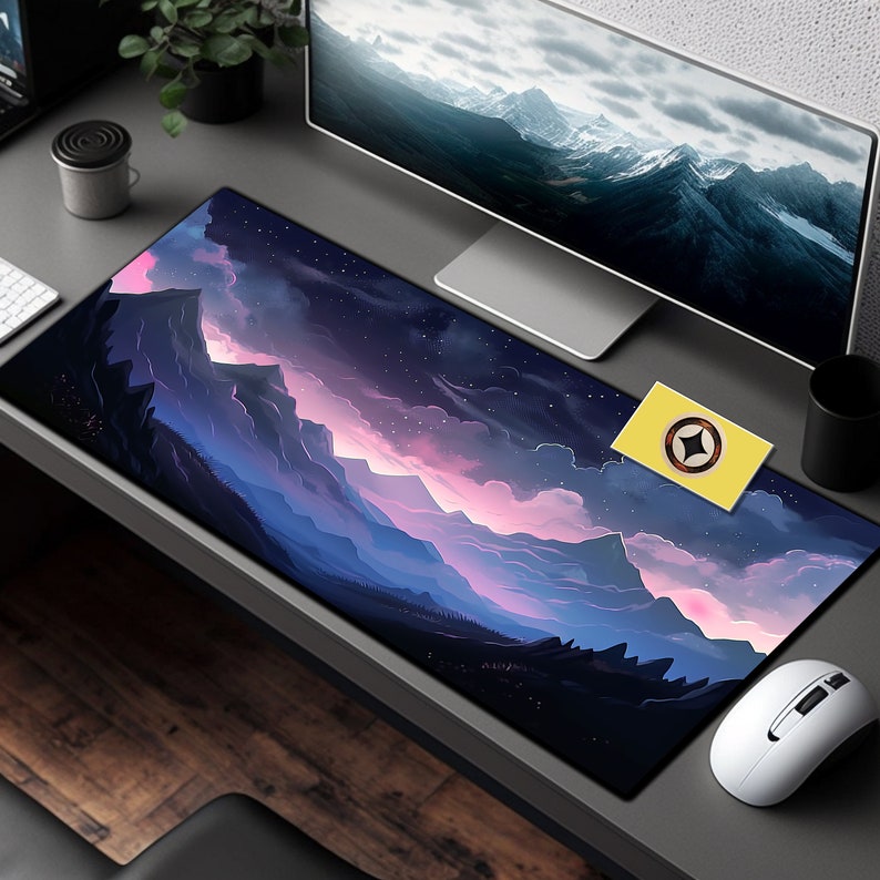 May include: A large black mouse pad with a purple and blue mountain range and starry sky design. The mouse pad is on a desk with a computer monitor, a yellow sticker, and a white mouse.