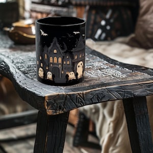 May include: Black ceramic mug with a spooky Halloween scene featuring a row of houses with glowing windows and ghosts. The mug is sitting on a dark wooden table.