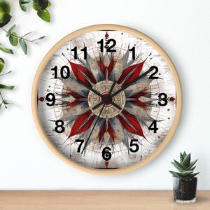 May include: A round wooden wall clock with a white face and black numbers. The clock face features a red and grey mandala design with a compass-like centre. The clock hands are black.