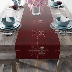 May include: A deep red table runner featuring a repeating pattern of crossed axes and geometric designs. The runner is laid on a wooden table, with place settings, wine glasses, and a vase of pink lilies. The overall theme is rustic and decorative.