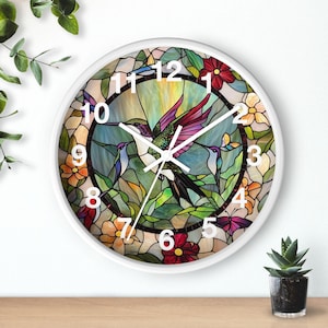 May include: White round wall clock with a stained glass design featuring a hummingbird, flowers, and leaves. The clock face has black Roman numeral hour markers.