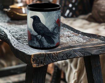 May include: Black ceramic mug featuring a detailed illustration of a raven perched on a branch with red roses, set against a backdrop of a full moon and a gray sky. The mug has a black interior and rim.