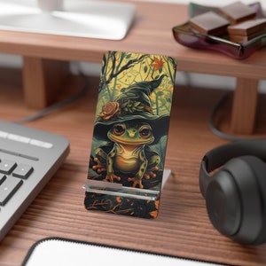 May include: A phone stand with a green frog wearing a witch's hat. The frog is sitting on a branch with leaves and flowers. The background is a forest scene with trees and vines.