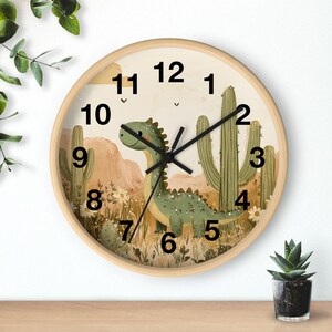 May include: A round wooden wall clock with a dinosaur and cactus design. The clock face has black numbers 1 through 12 and black hands. The background is a desert scene with a green dinosaur, brown mountains, and green cacti.