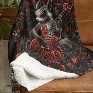 May include: A black and red throw blanket with a white sherpa backing features a stylized illustration of a furry creature playing an electric guitar. The creature is surrounded by ornate floral designs.