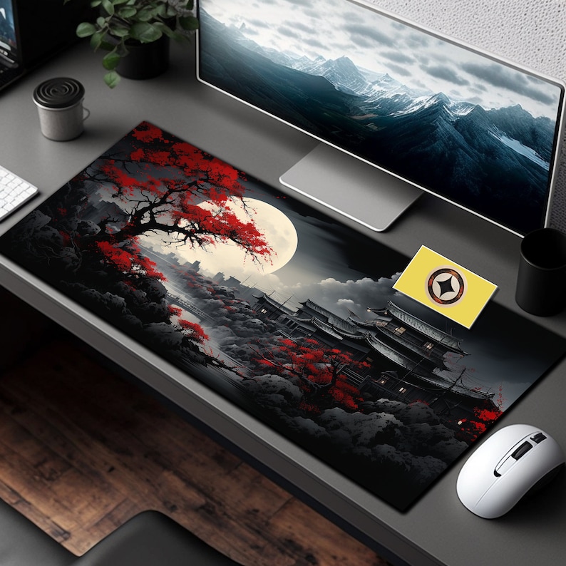 May include: Black and red mousepad with a Japanese-inspired design featuring a moon, trees, and a building. The mousepad is on a gray desk with a computer monitor, a mouse, and a small yellow card with a star on it.