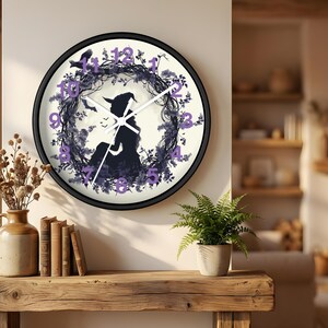 May include: A round wall clock with a black frame. The clock face features a silhouette of a witch with a hat, surrounded by a floral wreath and bats. The numbers are purple. The clock hands are white.