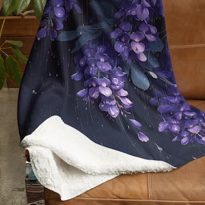 Puede incluir: Una manta suave y mullida con un fondo azul oscuro y un patrón floral de flores de glicinia moradas con gotas de agua. La manta está doblada y drapeada sobre un sofá de cuero marrón.