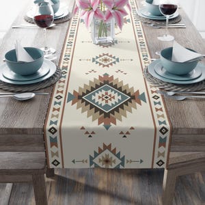May include: A cream-colored table runner with a geometric Southwestern design in shades of brown, teal, and tan. The runner is centered on a wooden table, with place settings and a vase of pink flowers visible.