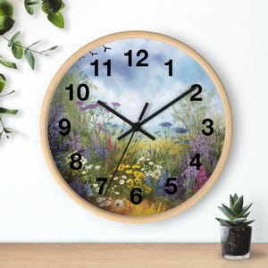 Könnte beinhalten: Runde Wanduhr mit hellem Holzrahmen. Das Zifferblatt zeigt eine farbenfrohe Wiesenlandschaft mit Wildblumen, blauem Himmel und schwarzen Zeigern. Die Zahlen sind schwarz.