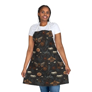 May include: An apron with a dark background featuring a repeating pattern of moths, flowers, and foliage in shades of brown, orange, and white. The apron has a white neck strap and waist ties. The apron is worn by a person.