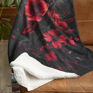 May include: A black and red fleece throw blanket with a floral pattern featuring large red flowers and a dark background. The blanket is folded and draped over a brown leather couch.