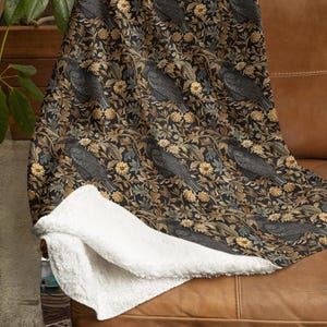 May include: A black and gold throw blanket with a repeating pattern of black birds and flowers. The blanket is made of a soft, plush material and has a white sherpa lining.