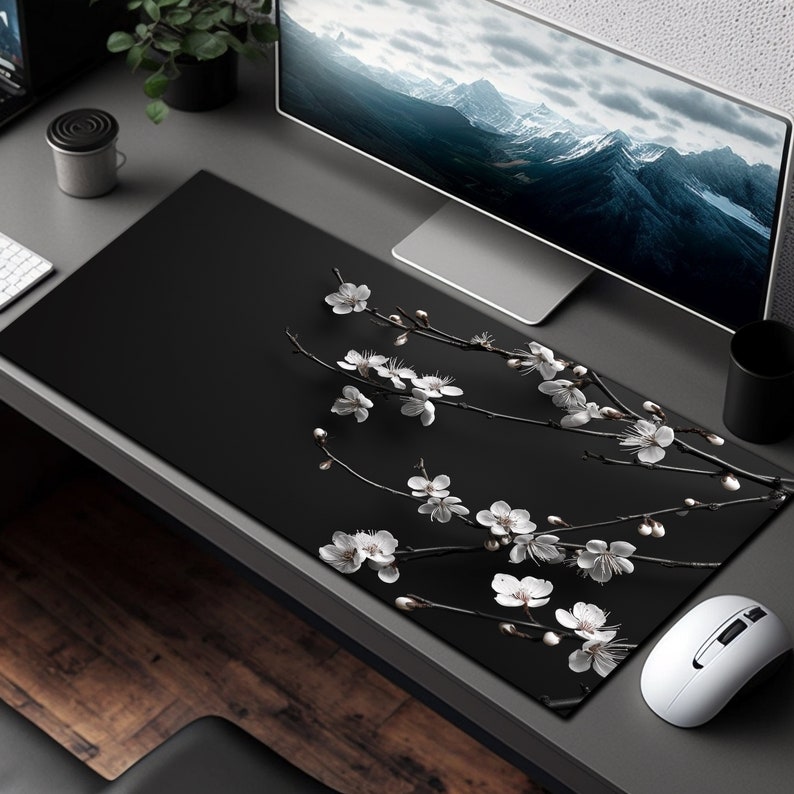 K&ouml;nnte beinhalten: Schwarze Schreibtischunterlage mit einem wei&szlig;en Blumendesign mit zarten Zweigen und Bl&uuml;ten. Die Unterlage liegt auf einem grauen Schreibtisch mit einem Computermonitor und einer Maus.