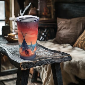 May include: A stainless steel tumbler with a colorful design of teepees against a sunset sky. The tumbler is sitting on a wooden table.