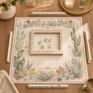 Könnte beinhalten: Ein komplettes Mahjong-Set mit einer beigen Spielfläche mit floralen und Kaktus-Designs. Das Set enthält Spielsteine, Leisten und einen Rahmen. Die Spielsteine sind weiß mit schwarzen und roten Symbolen. Das Set ist auf einem Holztisch angeordnet.