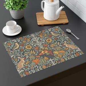 May include: A rectangular placemat featuring a dark blue background with a repeating pattern of brown rabbits, birds, sunflowers, wheat stalks, pumpkins, and a basket of fruit. The design includes intricate foliage in shades of green and orange.