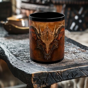 May include: A black coffee mug featuring a detailed illustration of a dragon's head. The dragon is depicted in shades of brown, orange, and gold against a textured background. The mug has a black rim and base, and is sitting on a dark wooden table.