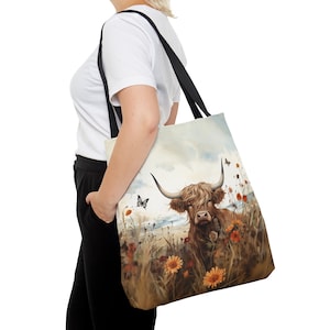 Könnte beinhalten: Eine braune und weiße Highland-Kuh mit langem, struppigem Fell steht auf einer Wildblumenwiese. Die Kuh schaut direkt in die Kamera. Der Hintergrund ist ein verschwommenes Bild von blauem Himmel und grünem Gras. Das Bild ist auf eine Canvas-Tragetasche mit schwarzen Henkeln gedruckt.