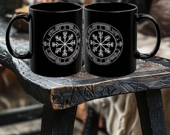 May include: Two black ceramic mugs with a white circular design featuring a snowflake-like symbol and runes. The mugs are displayed on a dark wooden surface, showcasing a rustic aesthetic.