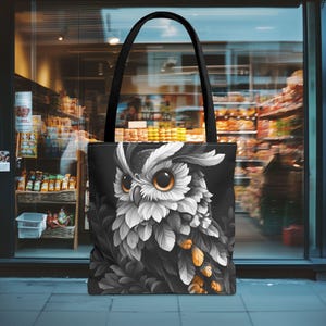 May include: Black tote bag with a detailed owl illustration in shades of grey, white, and gold. The owl has large orange eyes. The bag has black handles. The background is a blurred shop scene.