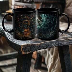 May include: Two black ceramic mugs with wraparound fantasy art. One mug features a tree with a house, the other a moonlit scene with a castle. Both mugs have black handles and are displayed on a wooden surface.