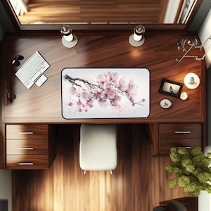 May include: A white desk mat with a watercolor painting of a pink cherry blossom branch. The mat is on a wooden desk with a keyboard, a lamp, and a chair.