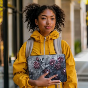 May include: A young person wearing a yellow hoodie and a backpack holds a laptop case with a floral design featuring dark red roses on a gray background.
