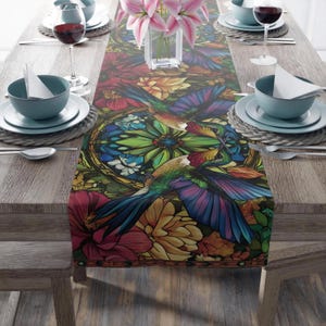 May include: A colorful table runner with a stained glass design featuring two hummingbirds flying among flowers. The runner has a dark green background with vibrant red, yellow, blue, and green accents.