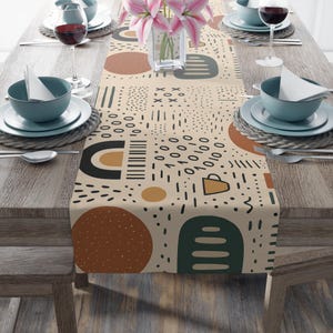 May include: A beige table runner with a repeating abstract pattern in black, brown, and green. The runner is decorated with circles, lines, and geometric shapes. The table is set with teal bowls, plates, and wine glasses.