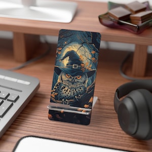 May include: A phone stand with a Halloween-themed design featuring an owl wearing a witch's hat. The owl is perched on a branch with leaves and a full moon in the background.