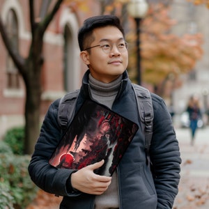 May include: A person wearing a black jacket and a grey turtleneck sweater is holding a laptop sleeve with a red and black landscape design. The sleeve features a red sun and a dark forest scene.