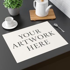 May include: A rectangular off-white placemat featuring the black text YOUR ARTWORK HERE centered on the surface. The placemat is positioned on a dark surface alongside a white ceramic cup and saucer, a white teapot on a wooden board, and a silver spoon.