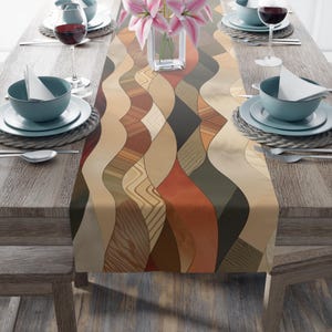 May include: A decorative table runner with a wavy, abstract pattern in shades of brown, beige, and rust. The runner is placed on a wooden table set with tableware, including plates, bowls, and wine glasses. A vase of pink flowers sits in the center.