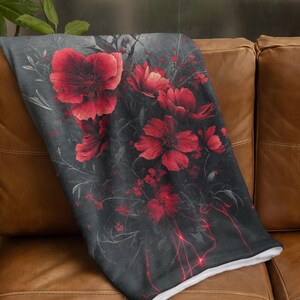 May include: A soft, plush throw blanket with a dark gray background and a floral design featuring red poppies. The blanket is folded and placed on a brown leather couch.