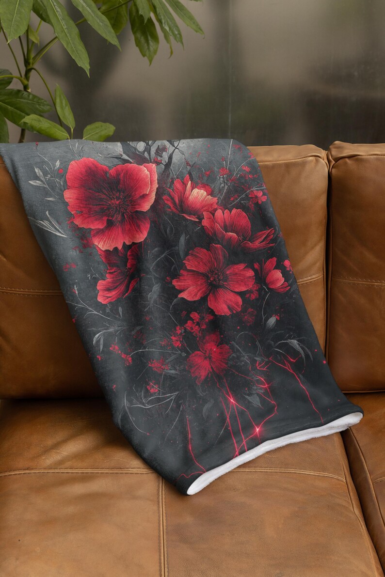 May include: A soft, plush throw blanket with a dark gray background and a floral design featuring red poppies. The blanket is folded and placed on a brown leather couch.