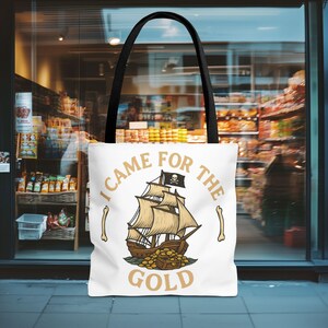 May include: White tote bag with black handles featuring a pirate ship illustration and the text "I came for the gold." The ship has tan sails and a black flag with a skull and crossbones. Gold coins are at the ship's base.