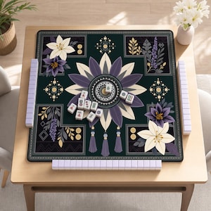 May include: A square mahjong mat featuring a dark green background with a central purple and cream floral mandala design, accented by corner botanical illustrations and tassels. White mahjong tiles are arranged on the mat and in racks.
