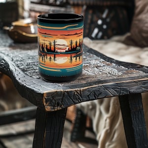 May include: A black ceramic mug with a colorful sunset scene and trees reflected in a lake. The mug is sitting on a rustic wooden table.